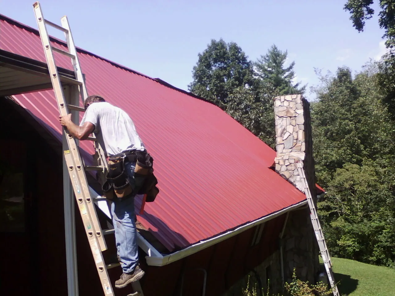 Roofer on a ladder beside a red metal roof and stone chimney — Soffit & Fascia Repair by Roofing Brothers in Canadensis